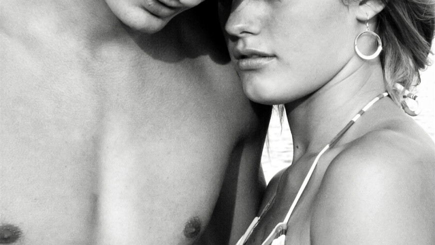 A young man and woman embrace, modeling in a beach scene facing the photographer