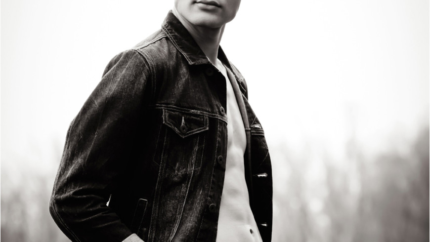 A young man models in a denim jacket and jeans while facing the photographer in front of distant trees in the Quad Cities