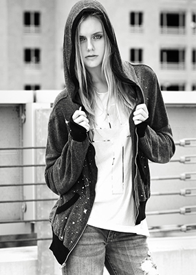 A young blonde woman in a black hoodie stands against an urban background facing the photographer in a white blouse and denim jeans