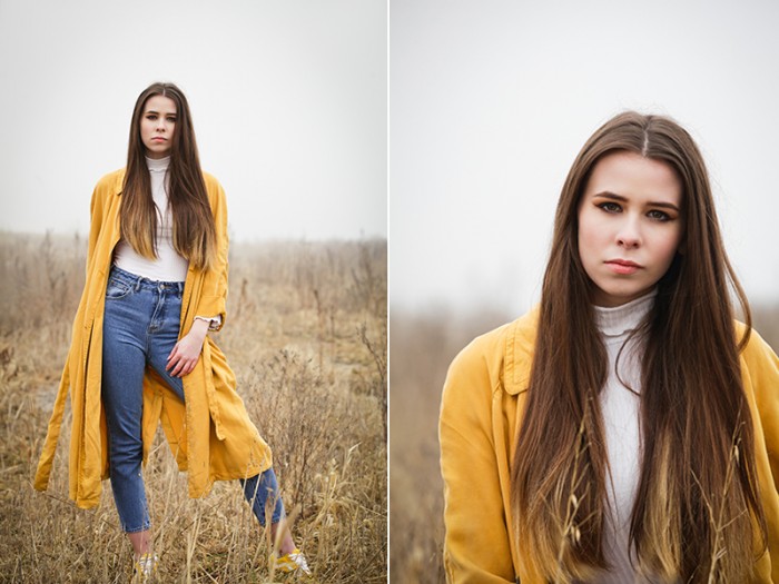 A young woman with long brown hair models in a grassy field facing the photographer while wearing a long yellow overcoat, a white top, and blue jeans