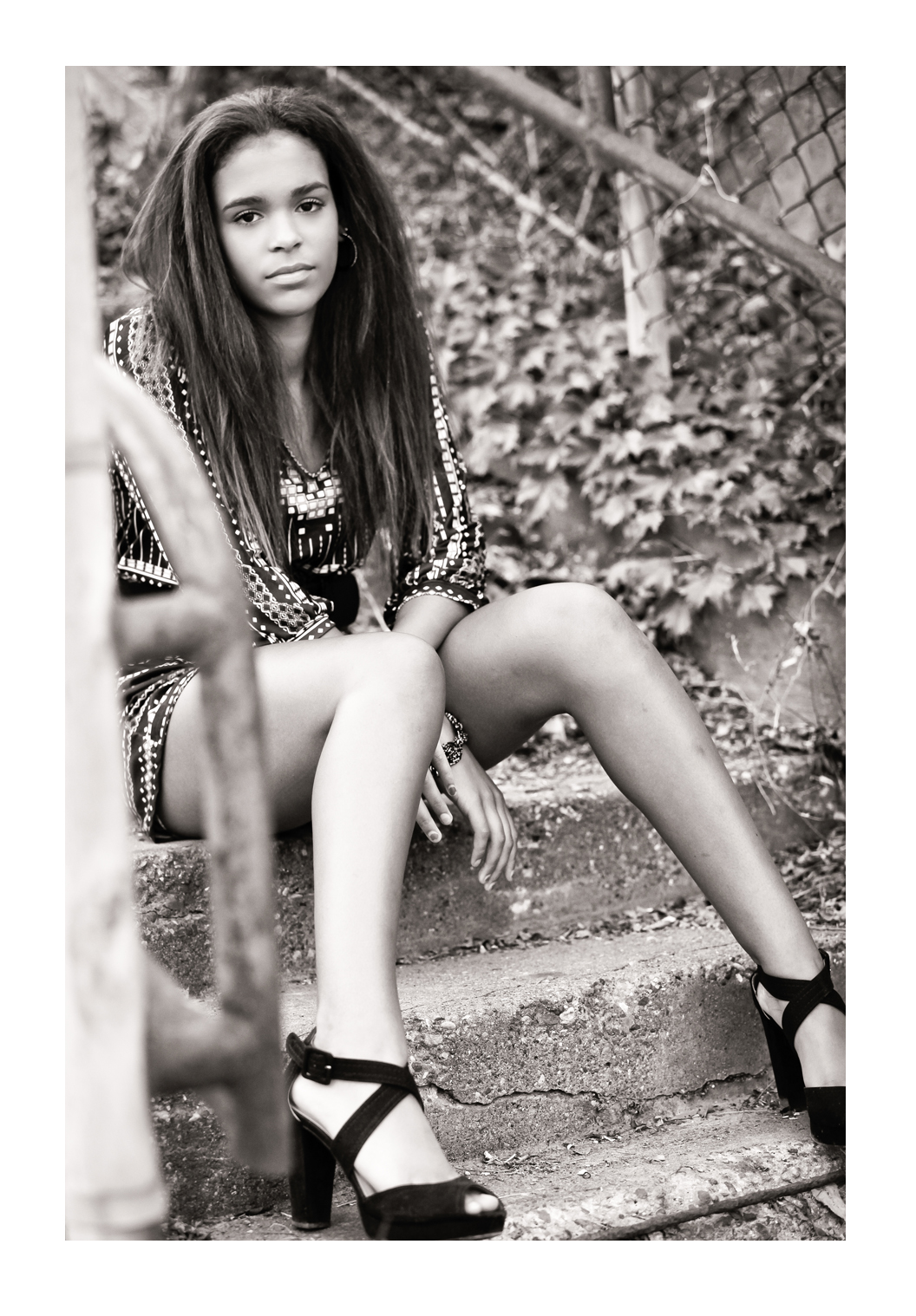 A young woman with straightened brown hair models facing the photographer while sitting on concrete steps wearing a glitzy dress and high heeled shoes