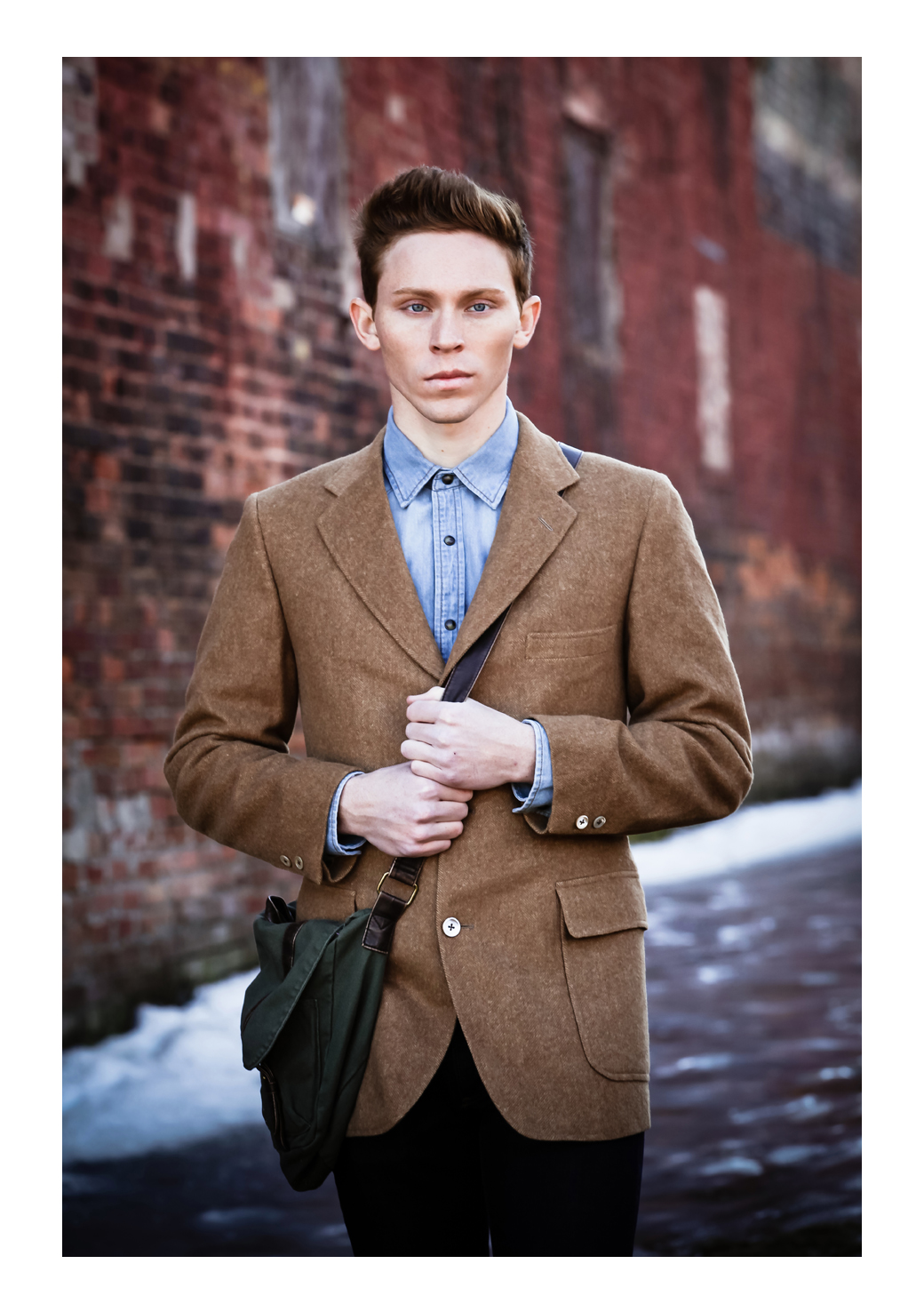 A young man with high auburn hair and freckles models, facing the cameraman while wearing a tweed suit in front of a brick wall