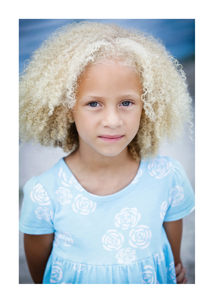 Young girl model with blonde ringlets stands facing the photographer in a blue flower-print dress