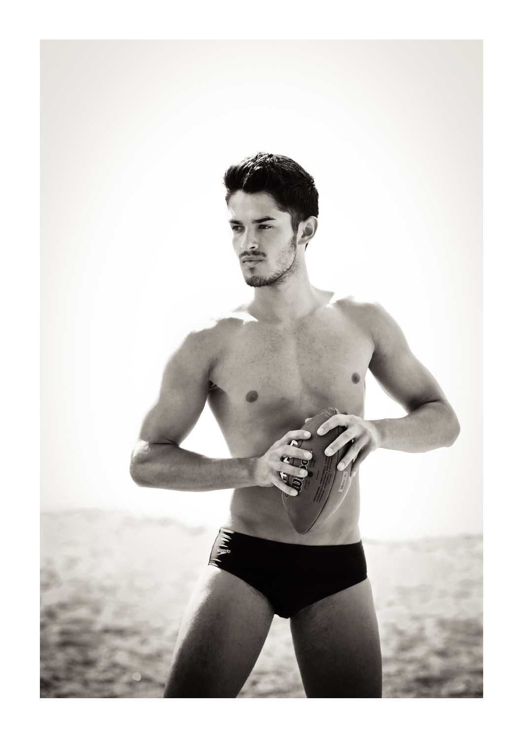 A young man with short dark hair stands in a black swim suit against a white sky holding a football in a ready-to-throw stance
