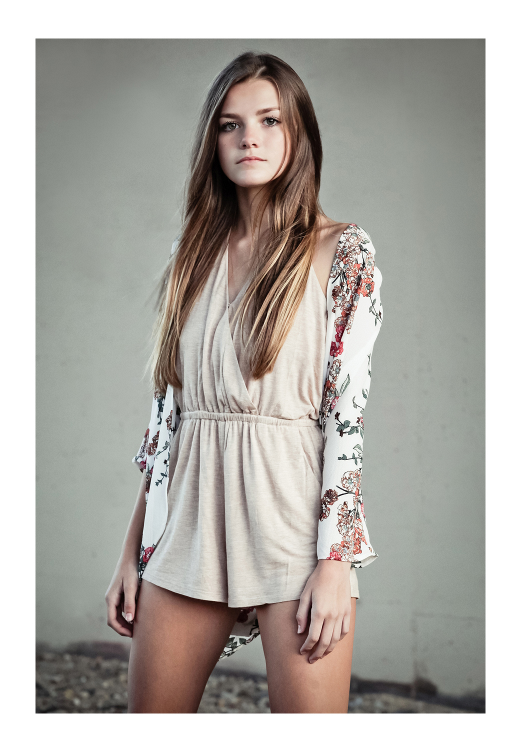 A young teen model with long brown hair stands facing the photographer wearing a canvas dress and a long flower-print blouse