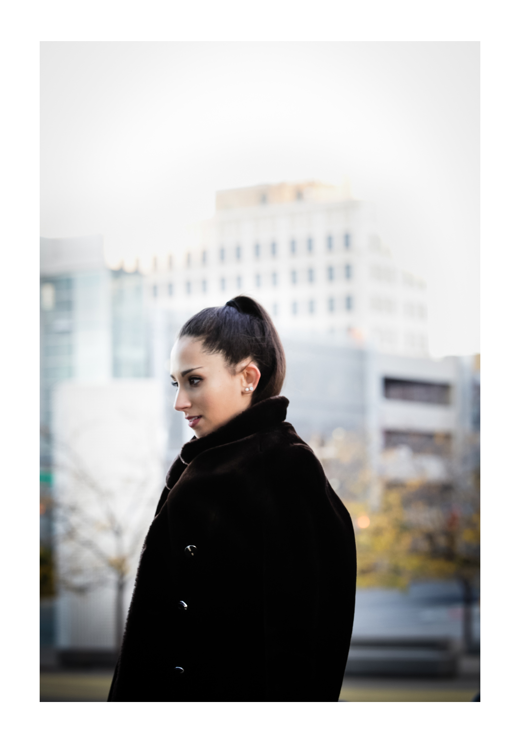 A model with dark brown hair in a pony tail wears a thick fur coat in front of a grey sky while gazing in the distance away from the photographer