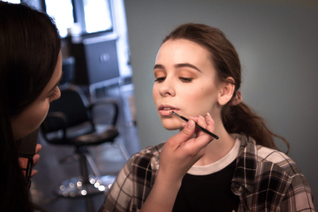 A young woman sits in a long-sleeved flannel shirt while an aesthetician applies makeup in a beauty salon
