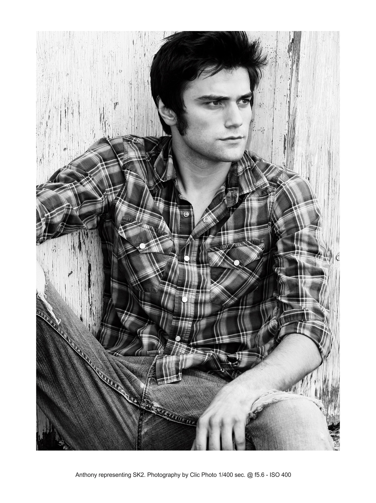 A young male model sits against a chipped paint wall with his hands on his knees. He wears a button-up shirt and jeans