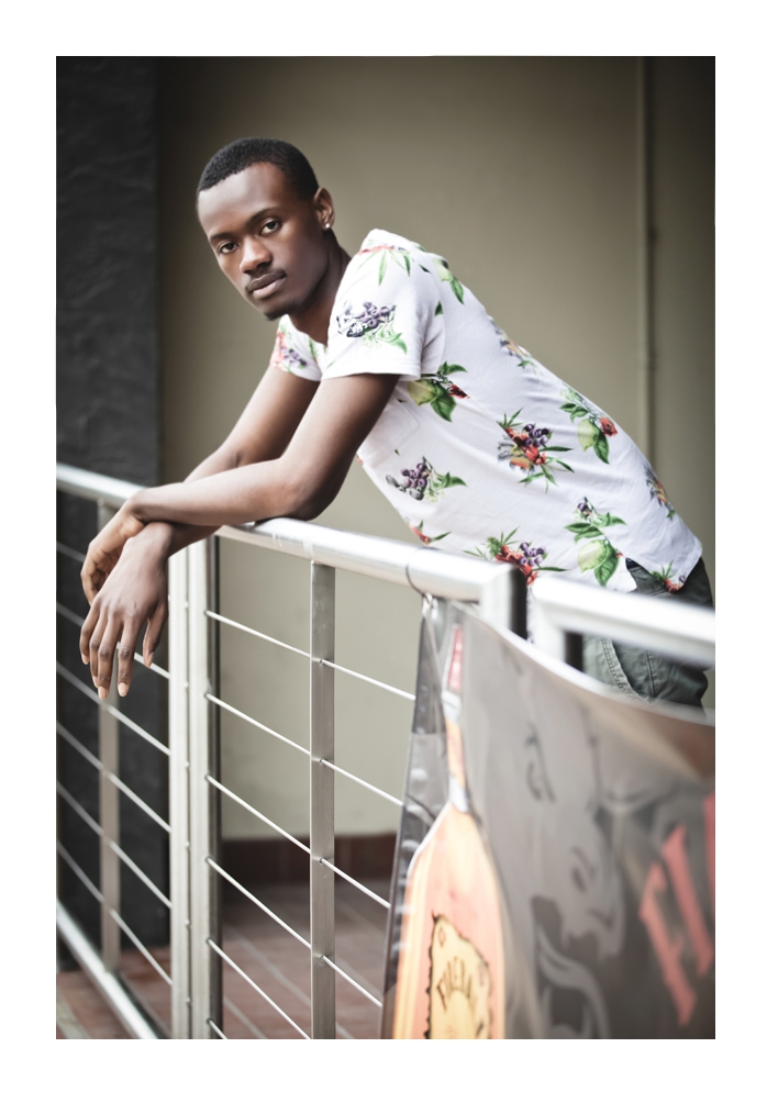 A young man models draped over a balcony railing facing toward the photographer while wearing a white t-shirt with a fruit basket pattern\