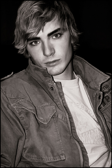 A young male model with side-swept dirty-blond hair in a darkened room sits facing the photographer while wearing a denim jacket