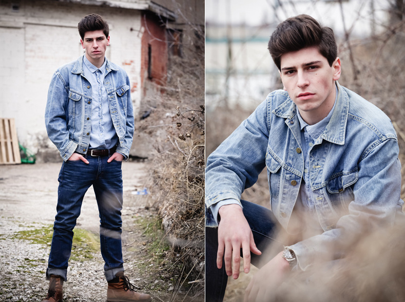 A young man models in a vacant lot filled with dried weeds. His dark brown hair is styled in a tall pompadour while he is wearing a denim jacket, a leather belt, and dark blue jeans