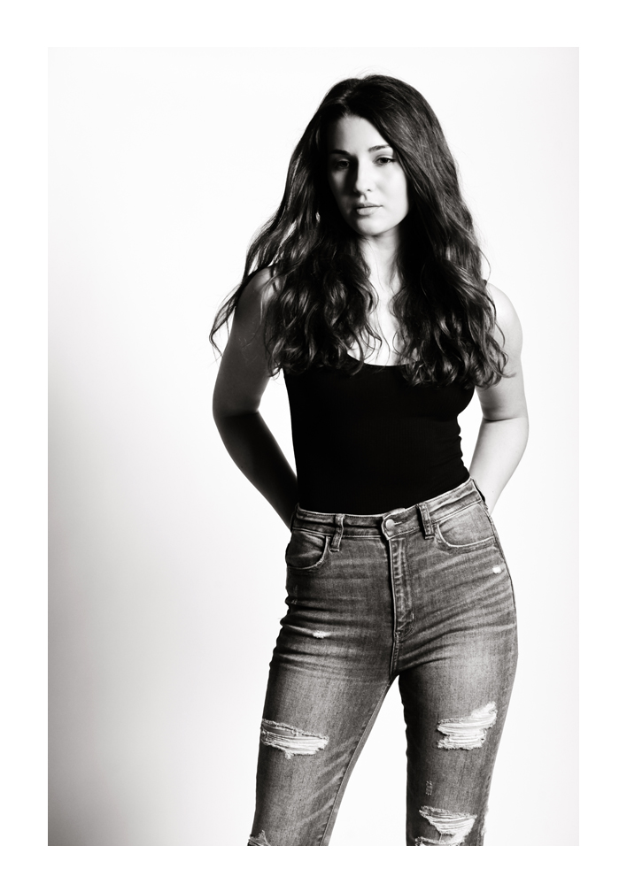 A young woman models, looking down with her dark hair flowing over her shoulders. A shadow crosses half of her face while she stands with her hands behind her back in a black top and denim jeans