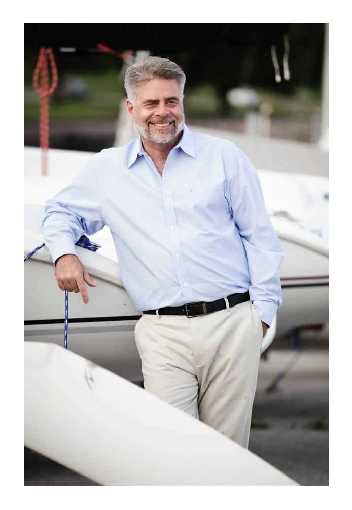 A middle-aged man with coiffed hair and a beard smiles, leaning at the front of his boat while looking into the distance