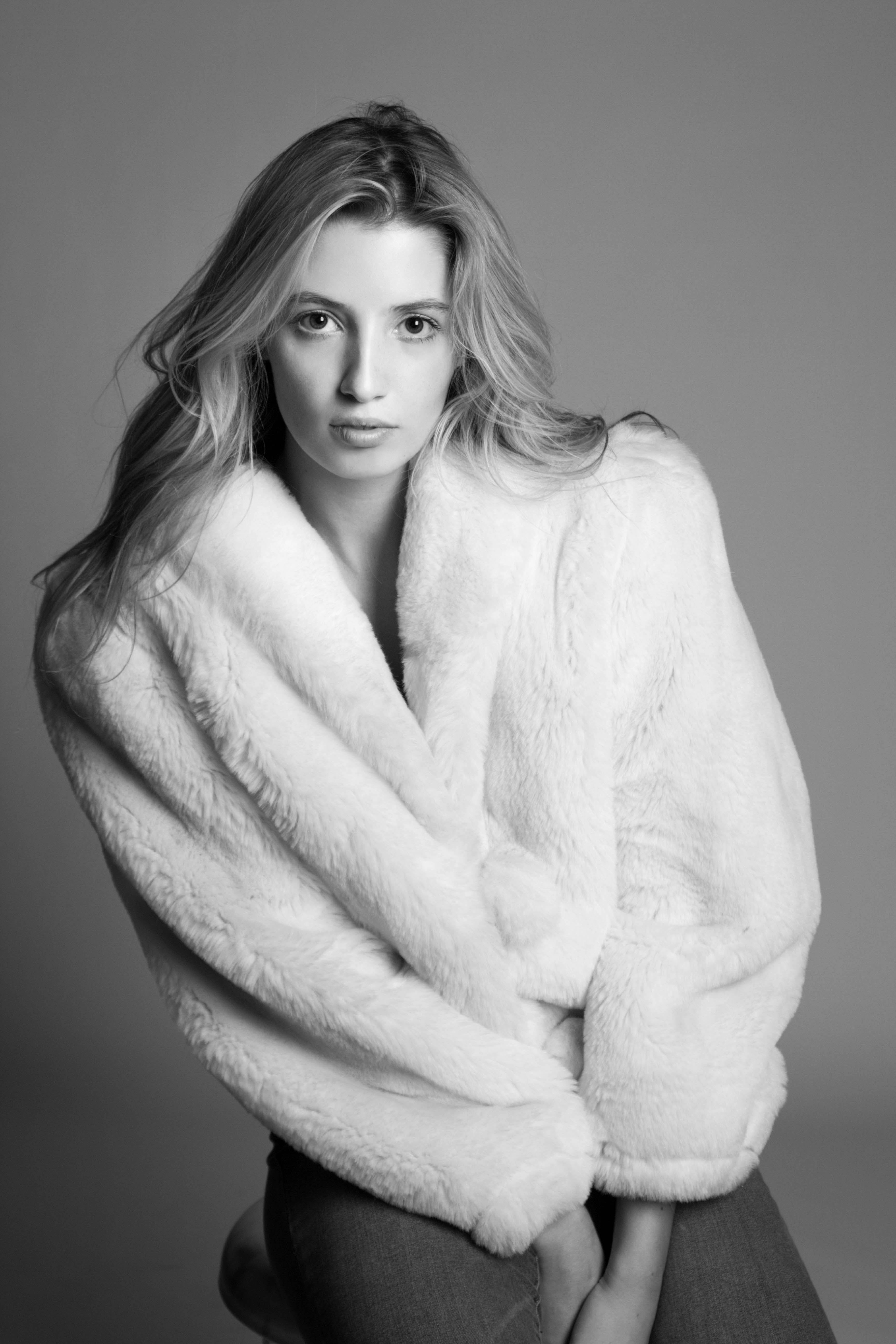 A greyscale photograph of a woman model sitting facing toward the photographer. She wears an oversized fleece jacket with her hands clasped