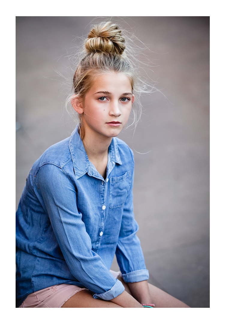 A young teen faces the photographer with a large top-knot in her blonde hair. She wears a blue denim button-up shirt and pink shorts