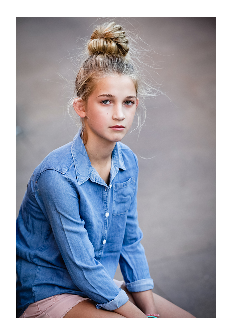 A young teen faces the photographer with a large top-knot in her blonde hair. She wears a blue denim button-up shirt and pink shorts
