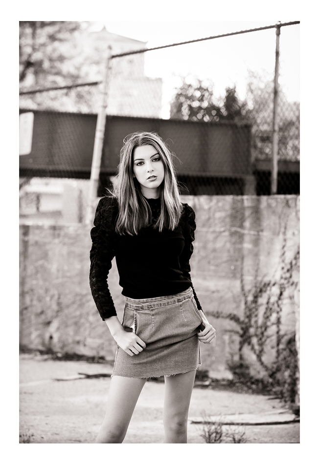 A brunette woman with hair past her shoulders models facing the photographer in front of a chain link fence. She wears a black, long-sleeved top and a denim skirt