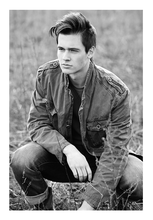A young model with a shiny black pompadour haircut crouches in the grass, looking away from the photographer toward the distance. He wears an autumn jacket and denim jeans