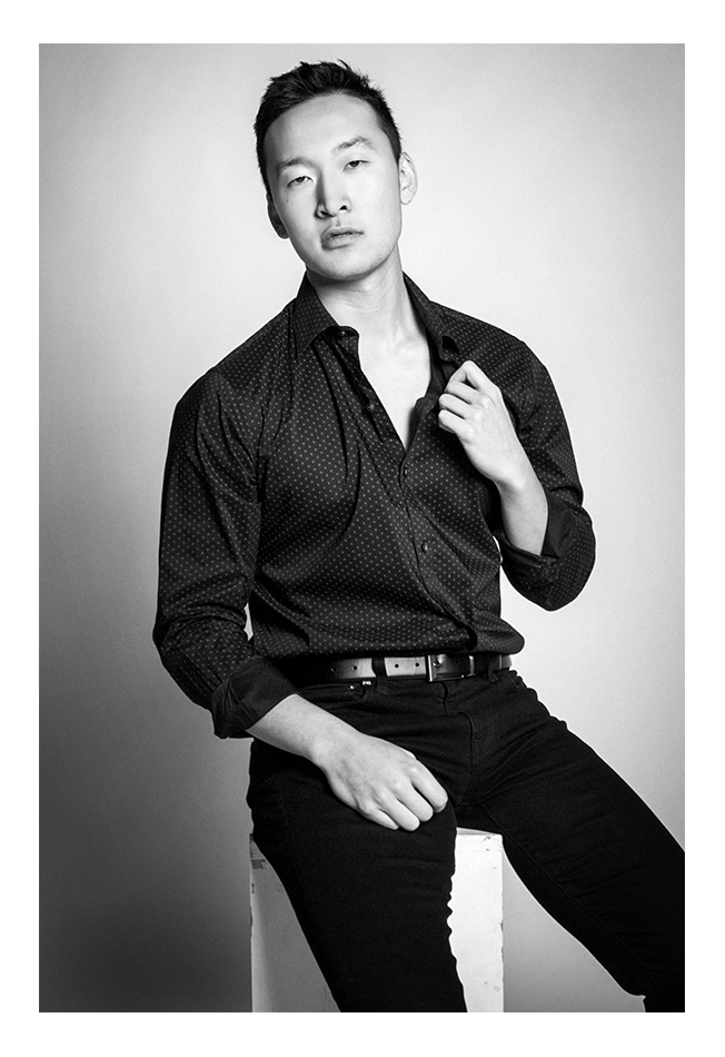 A black-haired man models facing toward the photographer. He sits in a photography studio wearing a dotted long-sleeved shirt and black pants with a belt