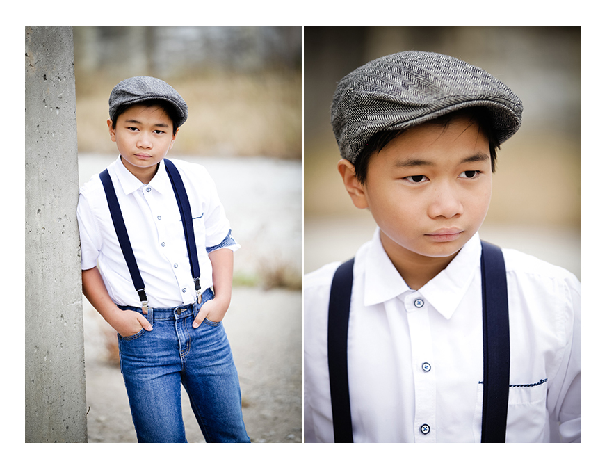 A young boy model leans against a cement pole, facing away from the photographer. He is wearing a grey vintage cap, a white button-down shirt with suspenders, and denim jeans