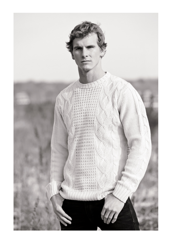 A young man models, looking in the distance. He has short light-brown hair and wears a white knitted sweater with elaborate diamond pattern with his thumbs looped in the pockets of his denim jeans