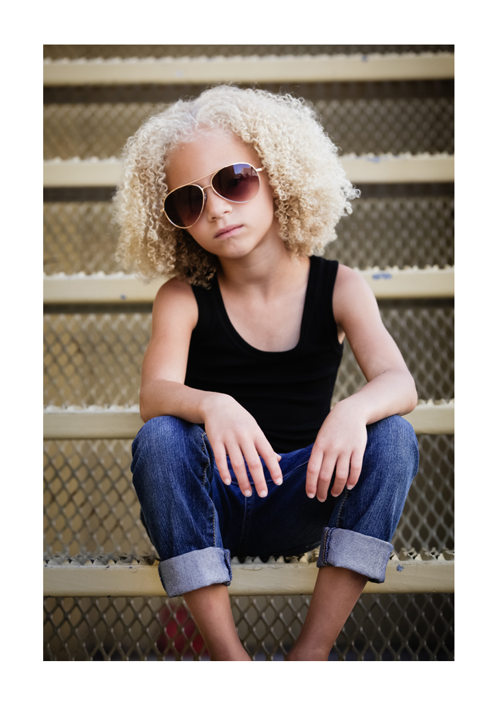 A young girl with blonde hair in tight ringlets models facing the photographer. She sits on corrugated steps wearing aviator sunglasses, a black top, and denim jeans