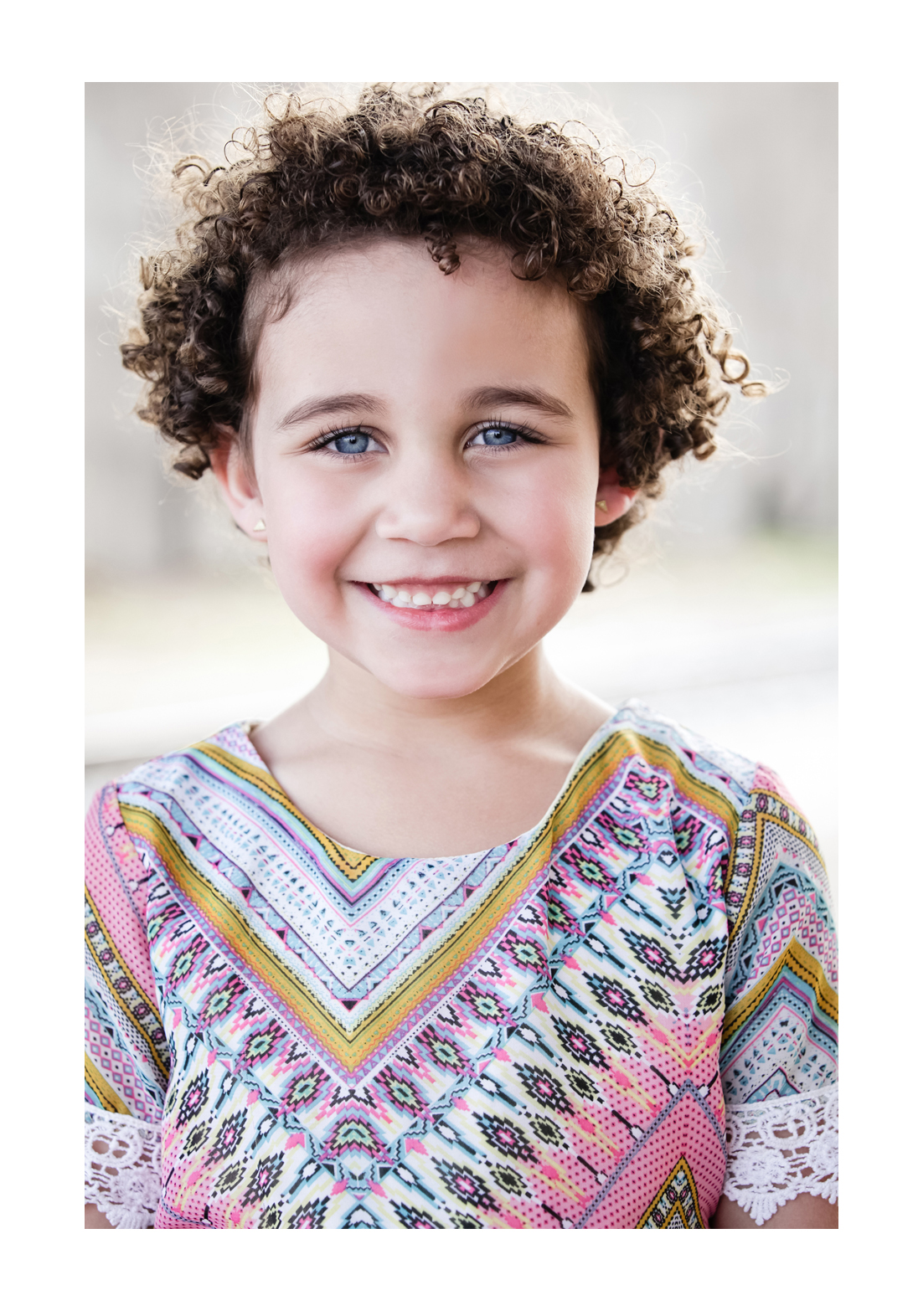 A young girl with curly brown hair faces the camera with a bright smile. She wears a colorful patterned shirt and two diamond earrings