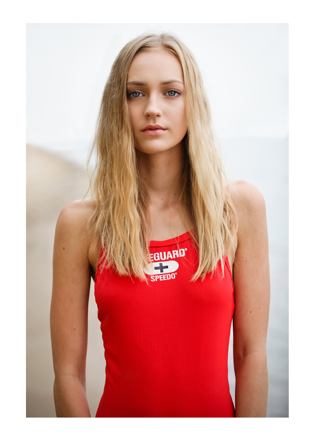 A young blonde woman wearing a red one-piece swim suit with the word Lifeguard printed on it stands facing the camera. Her toweled hair flows over her shoulders