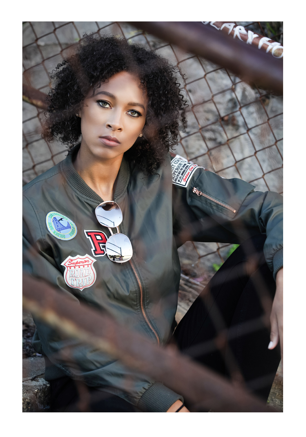 A teenage model sits behind a chain link fence, facing the photographer. She has curly black hair and wears a leather jacket with decorative patches