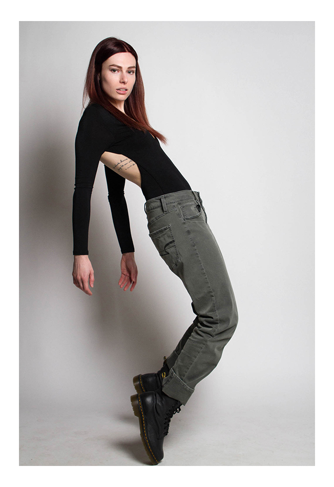 A woman with dark auburn hair models. She stands in an exaggerated backwards C pose facing the camera. She wears a black open-backed top with battleship grey pants and chunky black boots