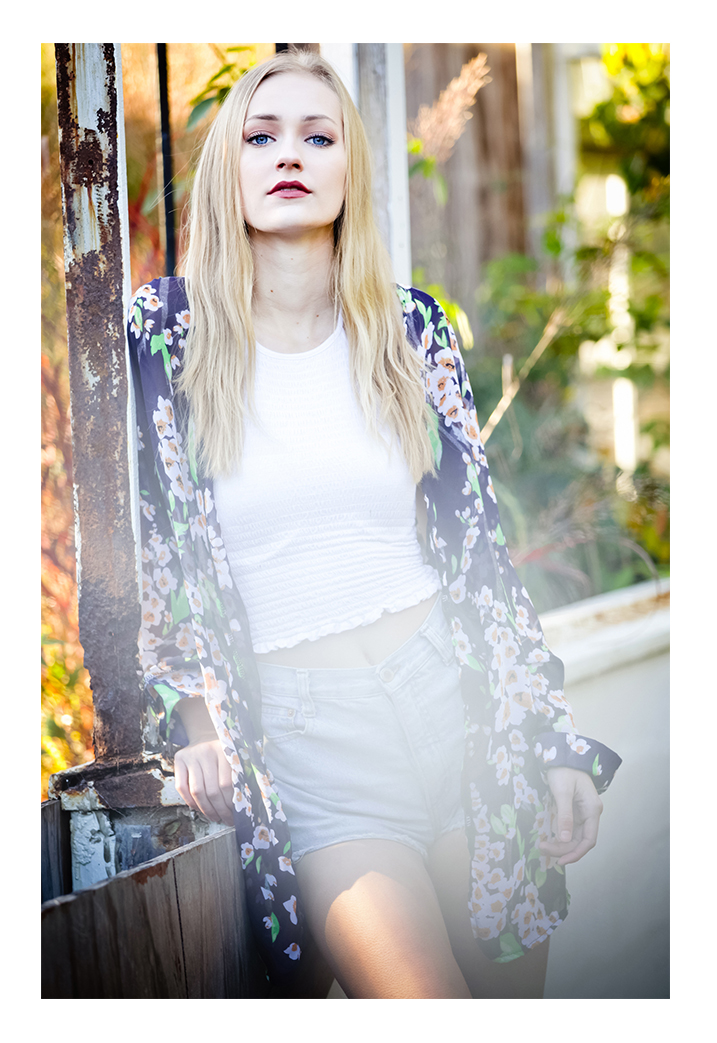 A blonde woman with long hair models, facing the photographer. She stands in an area with trees wearing a flowered love sleeve shirt, a white t-shirt and blue denim shorts