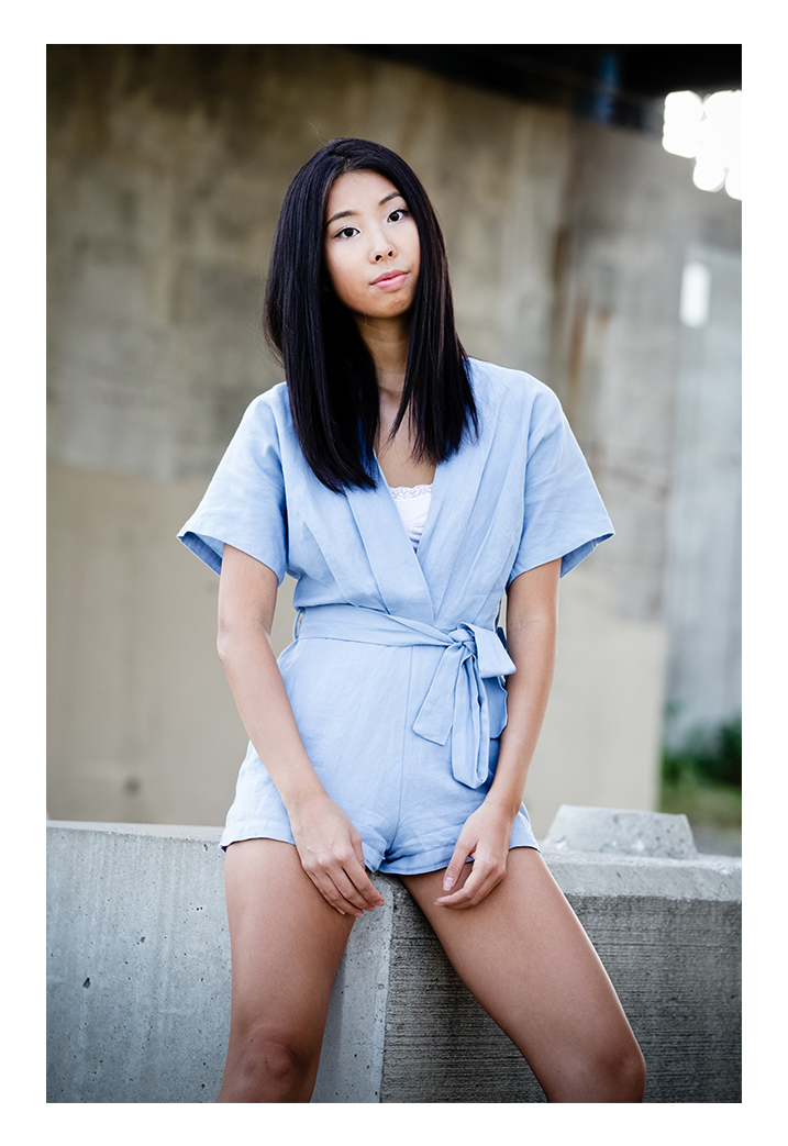 A woman models in front of a cement structure. She has long black hair and is wearing a periwinkle romper