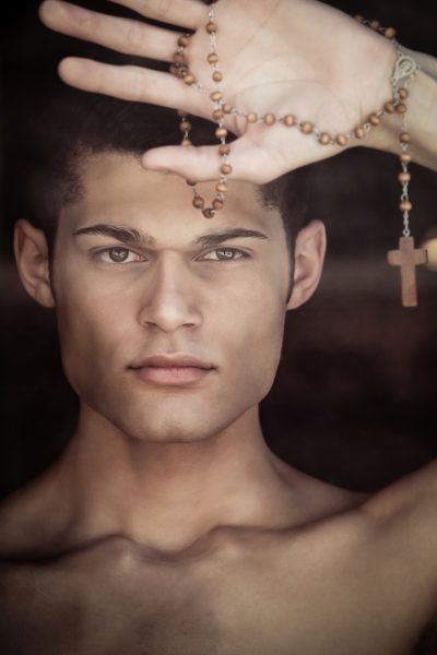 Young man with curly black hair models facing photographer holding up rosary beads
